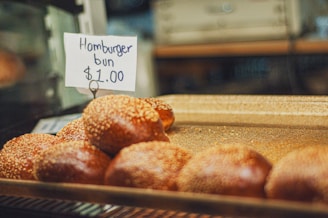 A tray of sesame-covered hamburger buns is displayed with a handwritten sign reading 'Hamburger bun $1.00.' The background appears to be a bakery or a deli with a glass display case.