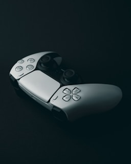 A game controller with a sleek design is placed on a dark surface, highlighting its ergonomic shape and button layout. The controller features a combination of smooth curves and well-defined buttons.