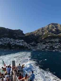 A group of people stand on the deck of a boat, taking photos as they travel across the water. The background reveals a picturesque coastal town nestled at the base of a mountain, with buildings densely packed along the shore and up the hillside. The sea is a rich blue, and the wake from the boat creates a white trail on the water. The scene is bathed in sunlight, highlighting the natural beauty of the location.