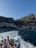 A group of people stand on the deck of a boat, taking photos as they travel across the water. The background reveals a picturesque coastal town nestled at the base of a mountain, with buildings densely packed along the shore and up the hillside. The sea is a rich blue, and the wake from the boat creates a white trail on the water. The scene is bathed in sunlight, highlighting the natural beauty of the location.