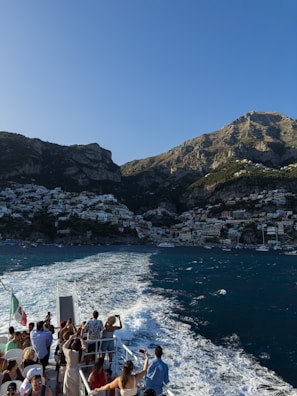 A group of people stand on the deck of a boat, taking photos as they travel across the water. The background reveals a picturesque coastal town nestled at the base of a mountain, with buildings densely packed along the shore and up the hillside. The sea is a rich blue, and the wake from the boat creates a white trail on the water. The scene is bathed in sunlight, highlighting the natural beauty of the location.