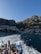 A group of people stand on the deck of a boat, taking photos as they travel across the water. The background reveals a picturesque coastal town nestled at the base of a mountain, with buildings densely packed along the shore and up the hillside. The sea is a rich blue, and the wake from the boat creates a white trail on the water. The scene is bathed in sunlight, highlighting the natural beauty of the location.