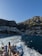 A group of people stand on the deck of a boat, taking photos as they travel across the water. The background reveals a picturesque coastal town nestled at the base of a mountain, with buildings densely packed along the shore and up the hillside. The sea is a rich blue, and the wake from the boat creates a white trail on the water. The scene is bathed in sunlight, highlighting the natural beauty of the location.