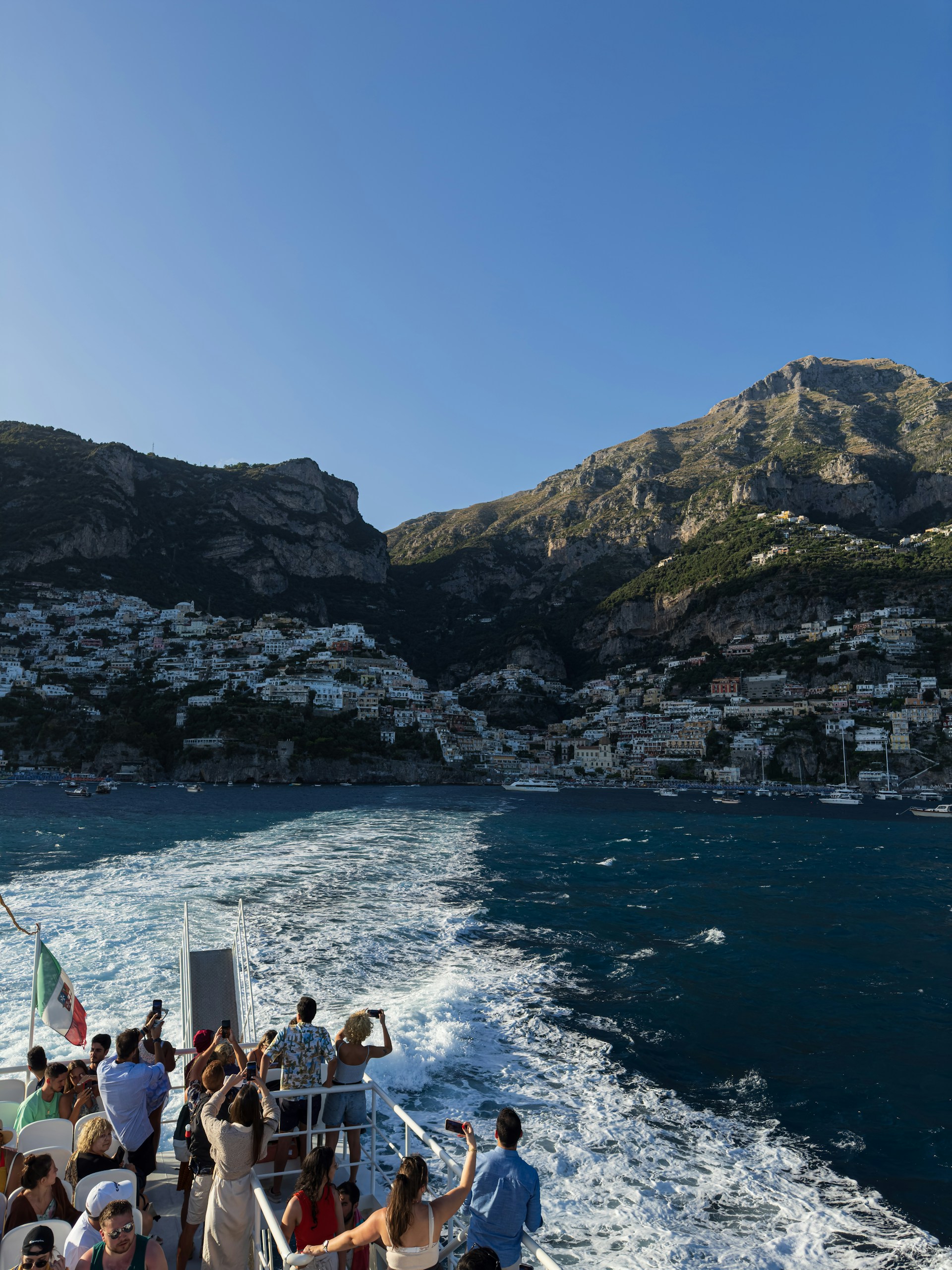 A group of people stand on the deck of a boat, taking photos as they travel across the water. The background reveals a picturesque coastal town nestled at the base of a mountain, with buildings densely packed along the shore and up the hillside. The sea is a rich blue, and the wake from the boat creates a white trail on the water. The scene is bathed in sunlight, highlighting the natural beauty of the location.