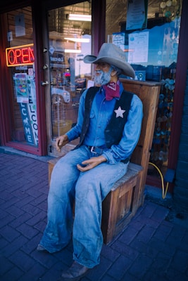 A mannequin dressed as an old western cowboy, complete with denim clothing, a cowboy hat, and a sheriff badge, is seated on a wooden bench. The figure is positioned in front of a storefront featuring an open neon sign and welcome signage, creating a rustic Wild West theme.