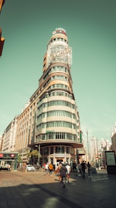 A tall, narrow building with a rounded facade and large signage on top stands at the corner of a bustling street. The architecture is characterized by a blend of modern and traditional elements, with large windows and a sleek design. People are walking on the pavement, some entering and exiting nearby shops. The scene is set against a clear sky, creating a vibrant urban atmosphere.