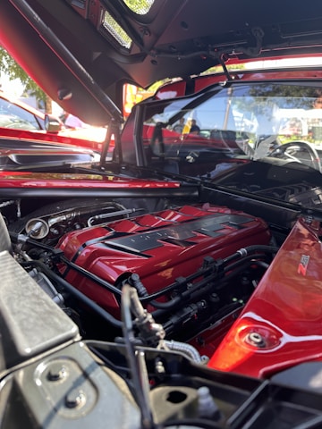 A close-up view of a car engine inside a red sports car, surrounded by metallic components and pipes. The engine cover is prominently colored in a shiny red with black accents, featuring visible branding. Sunlight highlights the polished surface, and parts of the exterior of the car can be seen in the background.