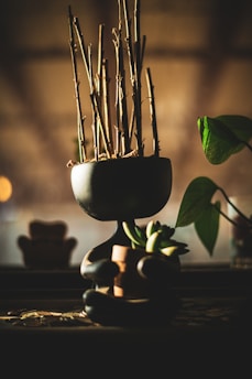 A minimalist black planter shaped like a seated human figure, holding a small pot with succulent plants. The planter contains dry stems, and a green leaf extends from the side. The background is softly blurred, giving a warm, cozy atmosphere.