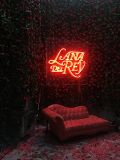 A neon sign displaying 'Lana Del Rey' glows prominently in red light. It is surrounded by a lush wall of red roses. In front of the sign is a plush, vintage-style red velvet couch, which is set against a backdrop of scattered rose petals on the floor, creating a romantic and theatrical atmosphere.