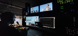 A person wearing headphones is sitting in a dark room, likely a control room or studio, in front of multiple screens and monitoring equipment. The screens display various images, including close-ups of two men in suits speaking, possibly during a broadcast or interview. There are colorful buttons and dials on the console, and several digital readouts with numbers.