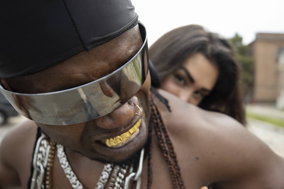 A person wearing large reflective sunglasses and a head covering, displaying gold jewelry including a grill and chains. In the background, there is another person partially visible, appearing over the shoulder.