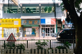 A bustling street scene featuring a row of shops and a property agency with bright signage, a pedestrian walking by, and a black vehicle passing along the road. In the foreground, a traffic sign and some greenery are visible.
