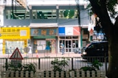 A bustling street scene featuring a row of shops and a property agency with bright signage, a pedestrian walking by, and a black vehicle passing along the road. In the foreground, a traffic sign and some greenery are visible.