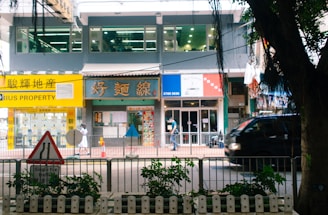 A bustling street scene featuring a row of shops and a property agency with bright signage, a pedestrian walking by, and a black vehicle passing along the road. In the foreground, a traffic sign and some greenery are visible.