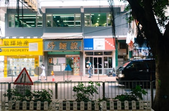 A bustling street scene featuring a row of shops and a property agency with bright signage, a pedestrian walking by, and a black vehicle passing along the road. In the foreground, a traffic sign and some greenery are visible.