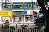 A bustling street scene featuring a row of shops and a property agency with bright signage, a pedestrian walking by, and a black vehicle passing along the road. In the foreground, a traffic sign and some greenery are visible.