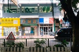 A bustling street scene featuring a row of shops and a property agency with bright signage, a pedestrian walking by, and a black vehicle passing along the road. In the foreground, a traffic sign and some greenery are visible.