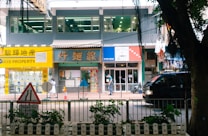 A bustling street scene featuring a row of shops and a property agency with bright signage, a pedestrian walking by, and a black vehicle passing along the road. In the foreground, a traffic sign and some greenery are visible.