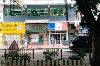 A bustling street scene featuring a row of shops and a property agency with bright signage, a pedestrian walking by, and a black vehicle passing along the road. In the foreground, a traffic sign and some greenery are visible.