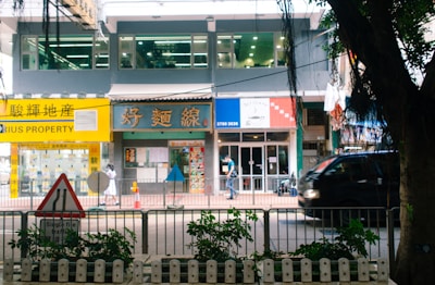 A bustling street scene featuring a row of shops and a property agency with bright signage, a pedestrian walking by, and a black vehicle passing along the road. In the foreground, a traffic sign and some greenery are visible.