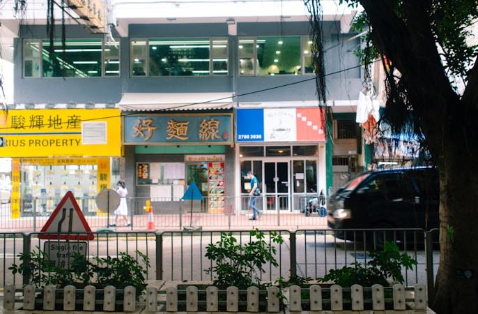 A bustling street scene featuring a row of shops and a property agency with bright signage, a pedestrian walking by, and a black vehicle passing along the road. In the foreground, a traffic sign and some greenery are visible.