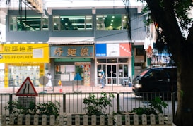 A bustling street scene featuring a row of shops and a property agency with bright signage, a pedestrian walking by, and a black vehicle passing along the road. In the foreground, a traffic sign and some greenery are visible.