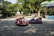 Two colorful bumper cars sit idle on a paved area in what appears to be an amusement park. The red bumper car features an animated face with a clown design, while the purple car is more minimalistic. Surrounding the area are lush green trees providing shade, and several colorful umbrellas are visible to the right.