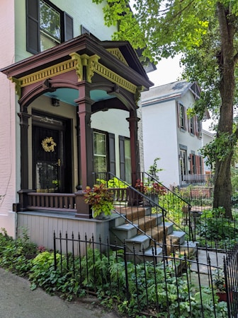 A charming house with a vintage porch adorned with a dark brown and green arch, surrounded by lush greenery. The porch features decorative support beams and potted plants with vibrant foliage. The house has light-colored brick walls and black shutters on the windows. A staircase with iron railings leads up to the entrance, which has a door with a decorative wreath. The setting is peaceful with a garden and trees.