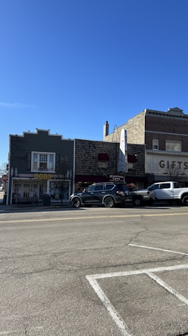 A street scene featuring several small buildings with shops and businesses. One of the shops has a sign with the name 'Bobs' and another named 'Treasure Cove'. Parked vehicles, including a black SUV and a white pickup truck, are visible along the street. The sky is clear and blue, suggesting pleasant weather.