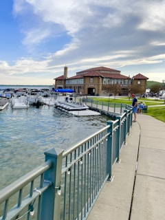 A waterfront scene with a large historic building labeled 'The Riviera' adjacent to a marina filled with several moored boats. A pathway with a sturdy metal railing runs along the water's edge, bordered by neatly maintained grass. The sky is partly cloudy with a mixture of blue and white, suggesting pleasant weather. There are a few people visible near the water and building, some sitting on the grass.