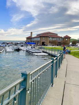 A waterfront scene with a large historic building labeled 'The Riviera' adjacent to a marina filled with several moored boats. A pathway with a sturdy metal railing runs along the water's edge, bordered by neatly maintained grass. The sky is partly cloudy with a mixture of blue and white, suggesting pleasant weather. There are a few people visible near the water and building, some sitting on the grass.