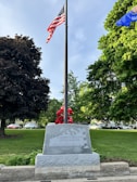 A flagpole with the American flag at the center, surrounded by lush green trees. A stone memorial at the base of the flagpole has an inscription dedicated to those who served in the military. Various red flowers are arranged around the memorial, and cars are visible in the background.