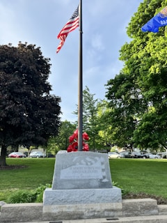 A flagpole with the American flag at the center, surrounded by lush green trees. A stone memorial at the base of the flagpole has an inscription dedicated to those who served in the military. Various red flowers are arranged around the memorial, and cars are visible in the background.
