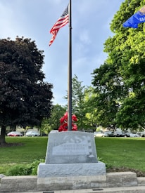A flagpole with the American flag at the center, surrounded by lush green trees. A stone memorial at the base of the flagpole has an inscription dedicated to those who served in the military. Various red flowers are arranged around the memorial, and cars are visible in the background.