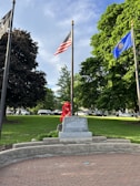 A memorial stone is prominently displayed in a park with lush green grass and large trees. The area is adorned with red flowers in front of the stone, and several flags are flying on tall poles. The American flag and a blue state flag are visible. Cars are parked on a road in the background.