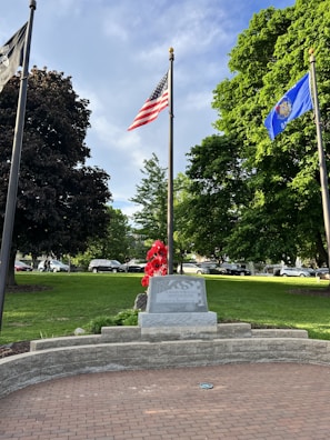 A memorial stone is prominently displayed in a park with lush green grass and large trees. The area is adorned with red flowers in front of the stone, and several flags are flying on tall poles. The American flag and a blue state flag are visible. Cars are parked on a road in the background.