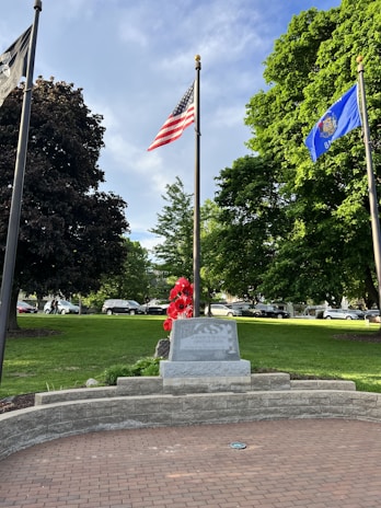 A memorial stone is prominently displayed in a park with lush green grass and large trees. The area is adorned with red flowers in front of the stone, and several flags are flying on tall poles. The American flag and a blue state flag are visible. Cars are parked on a road in the background.