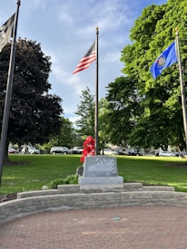 A memorial stone is prominently displayed in a park with lush green grass and large trees. The area is adorned with red flowers in front of the stone, and several flags are flying on tall poles. The American flag and a blue state flag are visible. Cars are parked on a road in the background.