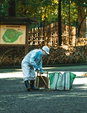 A person wearing a light blue uniform, cap, and face mask is cleaning the ground with a wooden tool. There is a large green bag next to them for collecting leaves on a gravel path in a forested area. A wooden signpost with a map and a bamboo fence are visible in the background among trees.