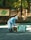A person wearing a light blue uniform, cap, and face mask is cleaning the ground with a wooden tool. There is a large green bag next to them for collecting leaves on a gravel path in a forested area. A wooden signpost with a map and a bamboo fence are visible in the background among trees.