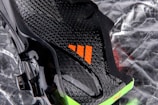 A close-up view of a sleek black sports shoe with a textured surface and an orange logo. The sole has a shiny finish with molded studs, and there is a neon green accent at the bottom.