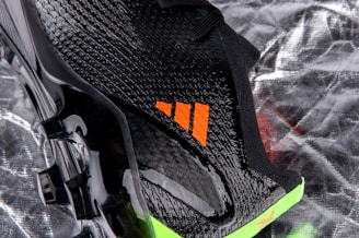 A close-up view of a sleek black sports shoe with a textured surface and an orange logo. The sole has a shiny finish with molded studs, and there is a neon green accent at the bottom.