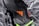 A close-up view of a sleek black sports shoe with a textured surface and an orange logo. The sole has a shiny finish with molded studs, and there is a neon green accent at the bottom.