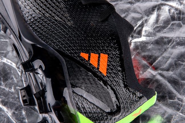 A close-up view of a sleek black sports shoe with a textured surface and an orange logo. The sole has a shiny finish with molded studs, and there is a neon green accent at the bottom.