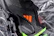 A close-up view of a sleek black sports shoe with a textured surface and an orange logo. The sole has a shiny finish with molded studs, and there is a neon green accent at the bottom.