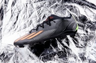 A black sports shoe with orange stripes and a sleek design lies on a crumpled, reflective silver surface. The shoe features textured fabric and a small green accent at the heel.