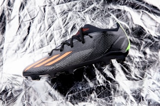 A black sports shoe with orange stripes and a sleek design lies on a crumpled, reflective silver surface. The shoe features textured fabric and a small green accent at the heel.