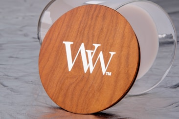 A wooden lid with a white 'WW' logo is positioned on a reflective surface. The lid appears to be resting against a transparent glass container.