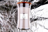 A tall glass candle with a wooden lid, labeled 'WoodWick' with the scent 'Sacred Smoke'. The background is a textured, crinkled metallic material.