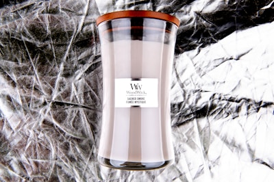 A tall glass candle with a wooden lid, labeled 'WoodWick' with the scent 'Sacred Smoke'. The background is a textured, crinkled metallic material.